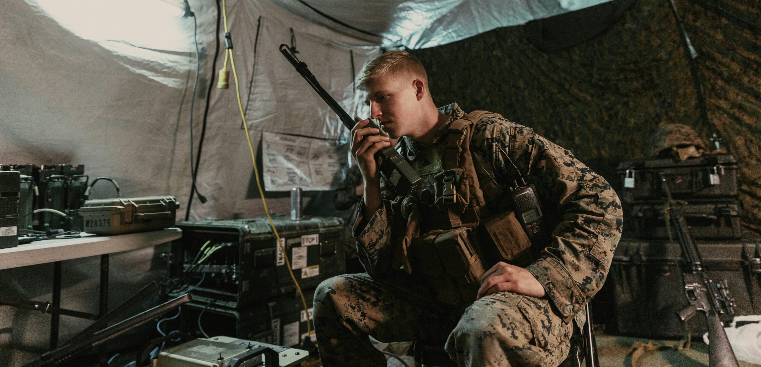 A member of the US Marine Corps using a handheld radio inside of a tent filled with communication equipment.