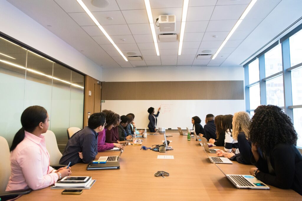 A group of people sitting around a conference table watching a woman pointing at a whiteboard.