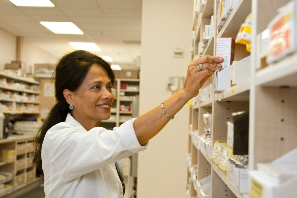 A pharmacist in a white coat reaching for a medication box on a shelf.